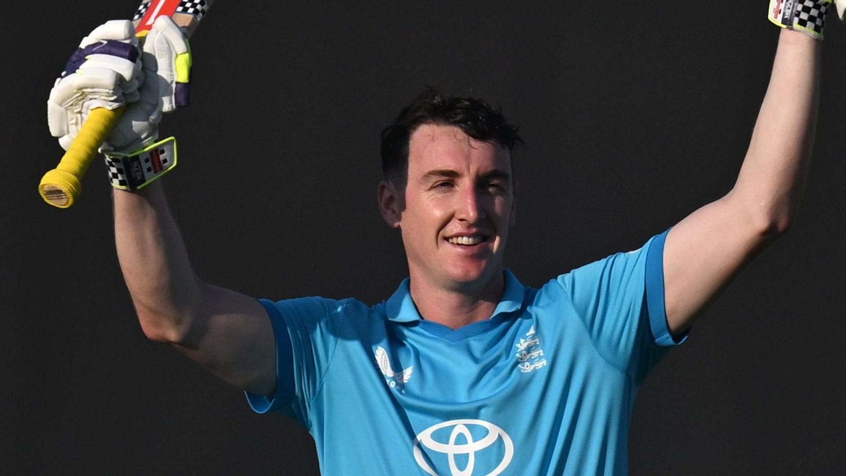 Harry Brook in a blue England kit. He is smiling with raised hands, holding his bat in one glove