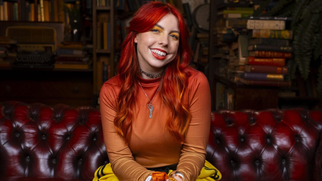 Ex-Traitors players talk tactics and celebrity final predictions A woman with bright red hair smiles t the camera. She is wearing an orange top and is sitting on a red sofa