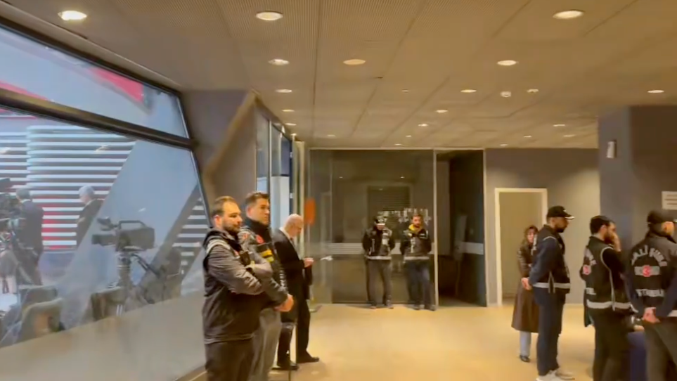 Police officers inside an office building with a TV studio visible through a window on the left of the frame.