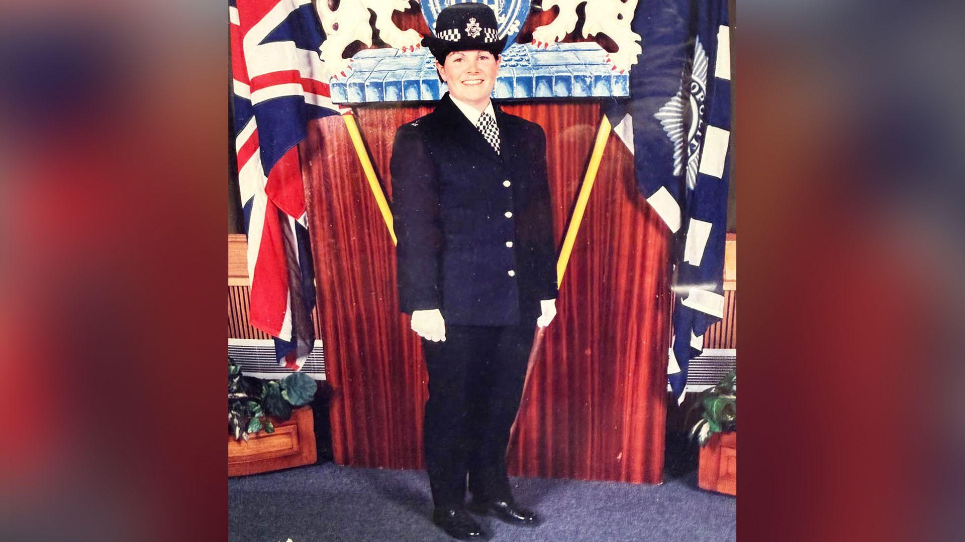 An official photo to mark when Linda xx joined the Metropolitan Police. Linda stands smiling into the camera wearing a full Met Police uniform and white gloves. She is flanked by a Union Jack flag on one side and a Met Police flag on the other.