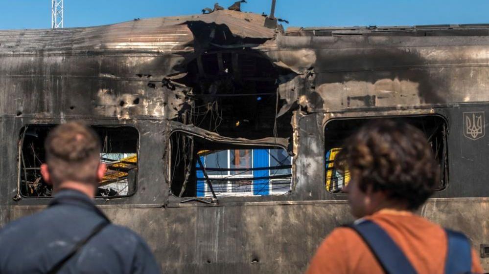 Two men stand in front of a burnt out train carriage