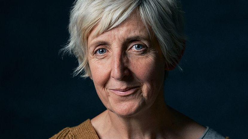 Close up of Julie Hesmondhalgh with short light-coloured hair looking directly at the camera. She is photographed from the shoulders up against a dark blue background. Soft lighting highlights her face. She is wearing a brown knit top layered over a light grey garment.