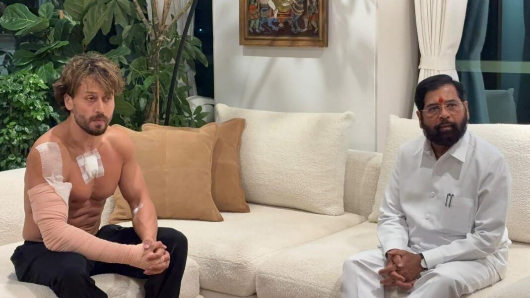 Two people sit on cream-coloured sofas in a living room during a meeting. One person on the left, Tiger Shroff, is shirtless and has bandages on the upper arm, shoulder, and chest, with a support wrap covering much of one arm. The person on the right, Eknath Shinde, wears a white outfit and glasses.