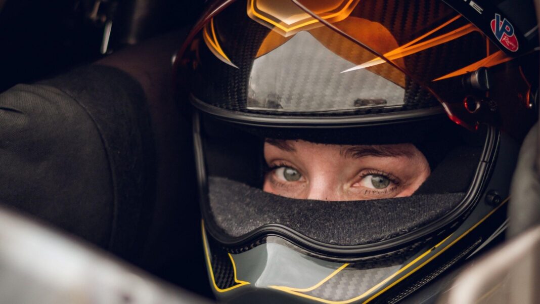 Swedish Top Fuel drag racing driver Susanne Callin wearing a black and orange racing helmet with just her eyes exposed through the visor, looking directly at the camera, taken at Mantorp in Sweden in 2025.