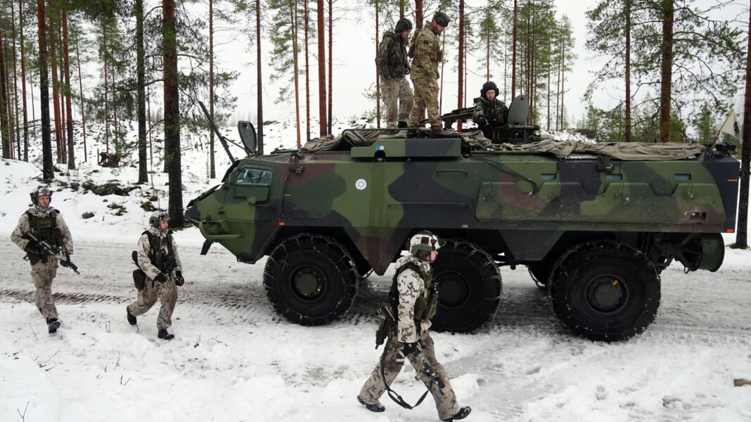 Three soldiers walk alongside an armoured tank in a snowy landscape. Three soldiers sit on top of the tank.