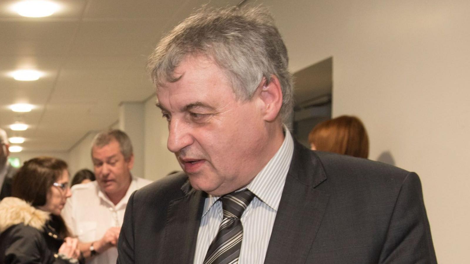 A man with grey hair looks to the side, he has a black suit an stiped shirt with a black and grey tie