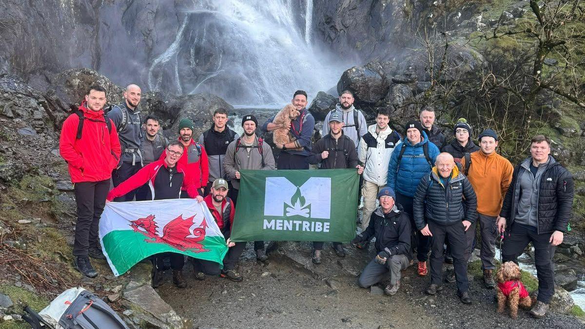 A large group of hikers pose at the base of a waterfall cascading down a steep rocky cliff.  The group stands on a rocky path surrounded by mossy rocks and bare trees, with several people holding a Welsh flag and a green “MENTRIBE” banner. Two dogs are visible, one being held by a participant and another wearing a red coat near the front. Backpacks rest on rocks in the foreground, while mist from the waterfall rises behind the group in an outdoor mountain setting.