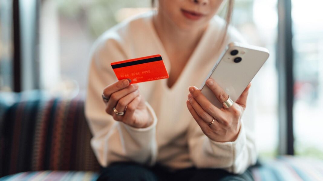 A woman sitting on a sofa and shopping online with her white phone while holding her red bank card in the other hand. She is leaning forward and wearing dark trousers and a white jumper.