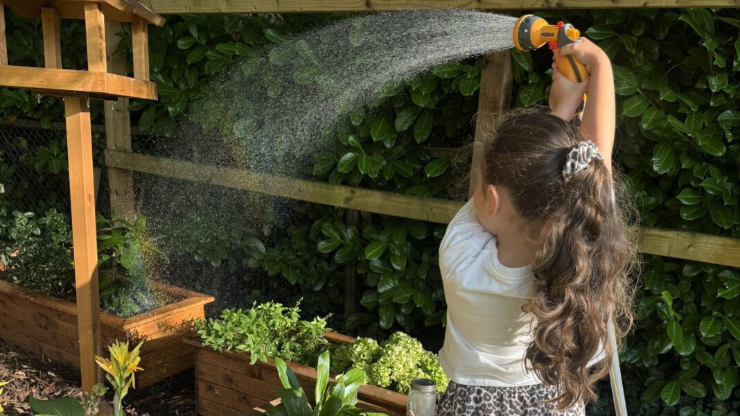 Child standing in a garden in the foreground, holding a hose spray nozzle above their head and watering raised wooden planters. Water arcs from the nozzle across the planters, which contain various flowers and leafy plants. A wooden bird-table-style structure stands beside the planters. A wooden fence and dense hedging form the background.