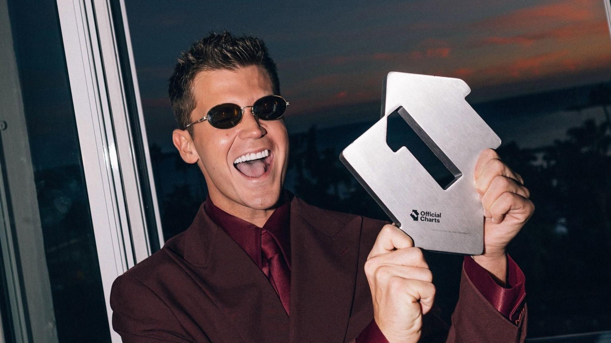 Jamie Miller in a red suit wearing sunglasses. He is smiling and holding an award for the UK number one single.