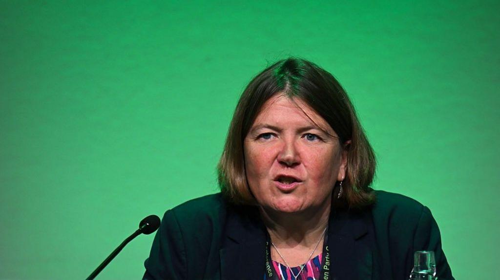 Ellie Chows in front a green background. She is speaking into a microphone, there is also a glass bottle of water on the desk she sits behind.