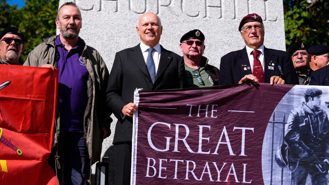 Sir Iain Duncan Smith stands with Northern Ireland veterans as they stage a protest in front of a statue of Winston Churchill outside the Houses of Parliament in London in September 2026. Smith is helping veterans in berets to hold up a banner which depicts a image of soldier on patrol holding a gun beside the slogan 