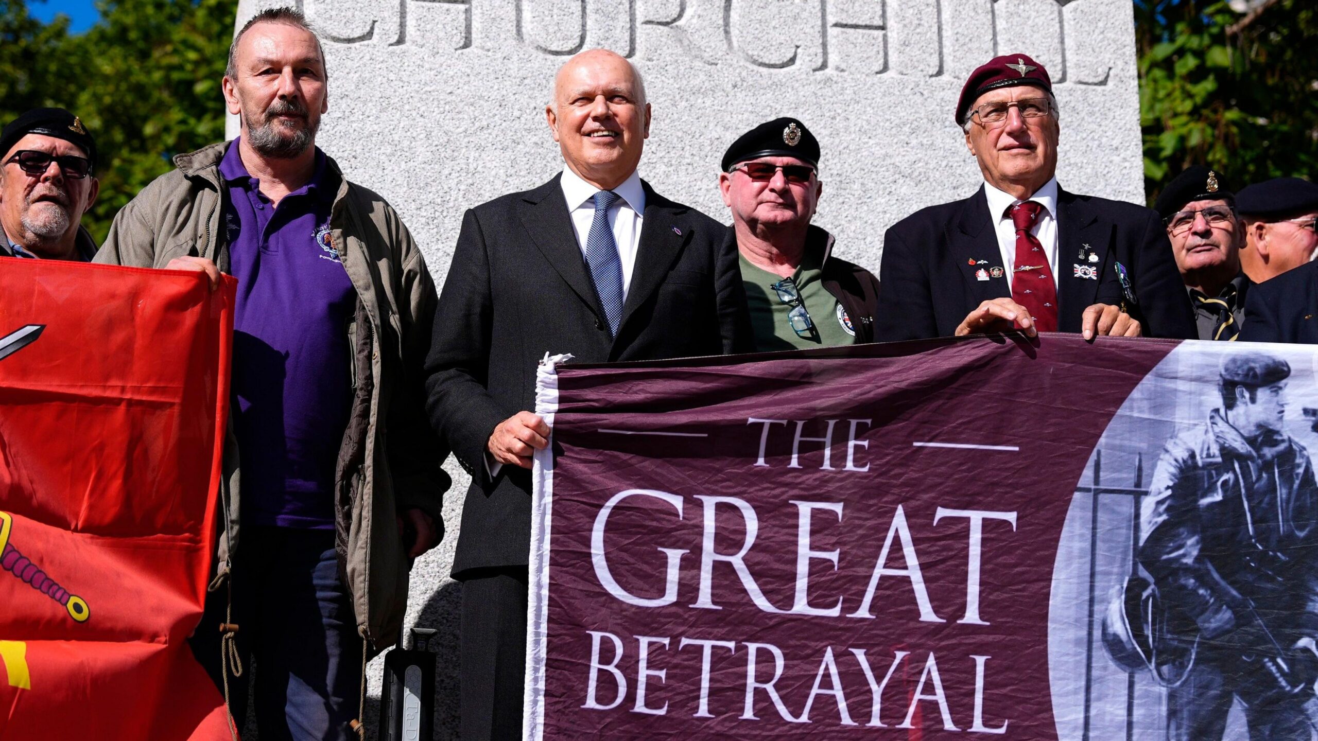 Sir Iain Duncan Smith stands with Northern Ireland veterans as they stage a protest in front of a statue of Winston Churchill outside the Houses of Parliament in London in September 2026.  Smith is helping veterans in berets to hold up a banner which depicts a image of soldier on patrol holding a gun beside the slogan "The Great Betrayal".