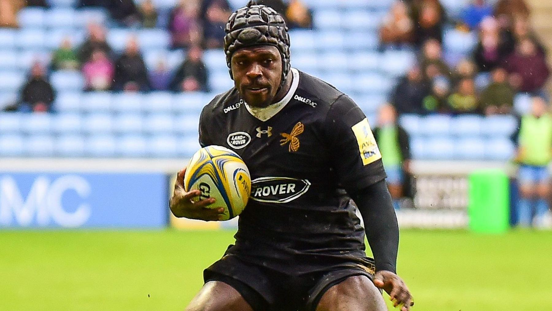 Christian Wade playing for Wasps in their all-black kit, with the rugby ball tucked under his right arm and switching weight from his right to his left side
