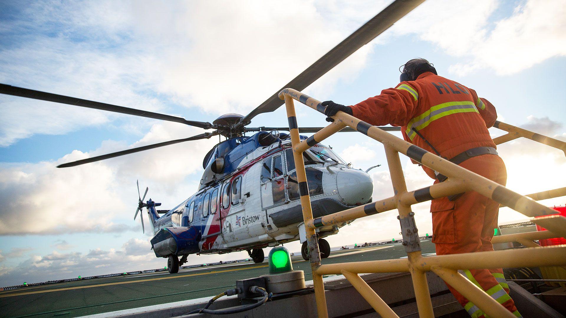 A helicopter lands on an oil rig and a man in orange overalls is heading towards it