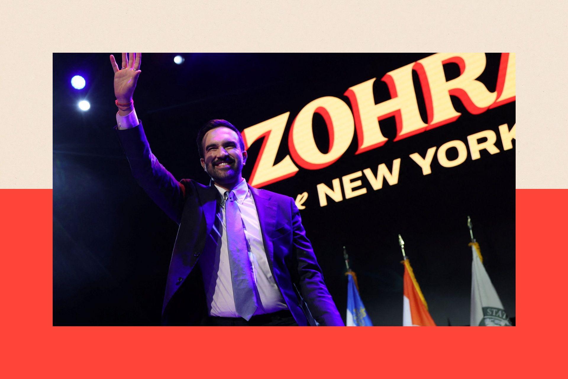 Zohran Mamdani stands on stage waving under a sign saying 'Zohran for New York'