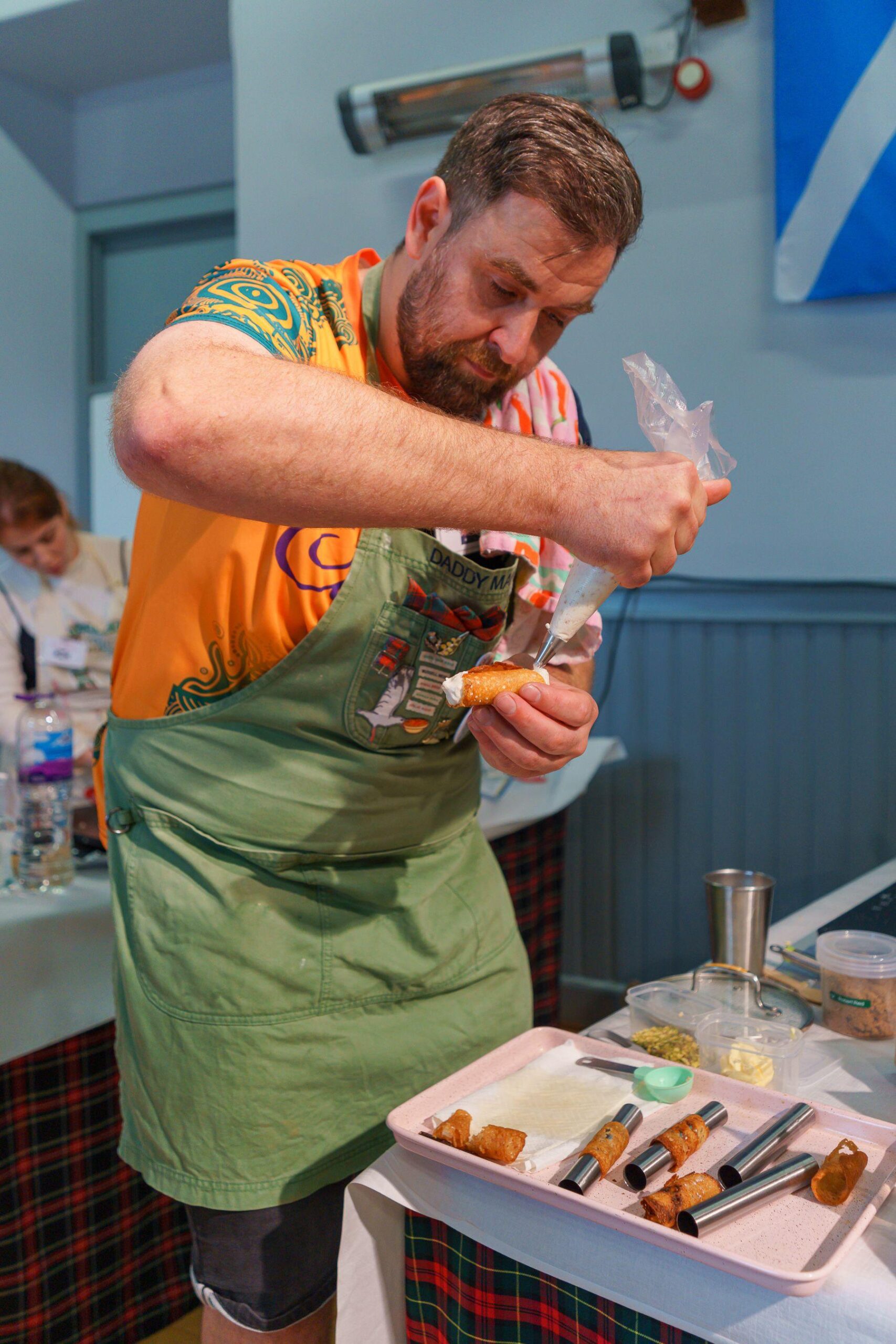 A bearded man in an orange T-shirt and green apron fills some round pastries with cream