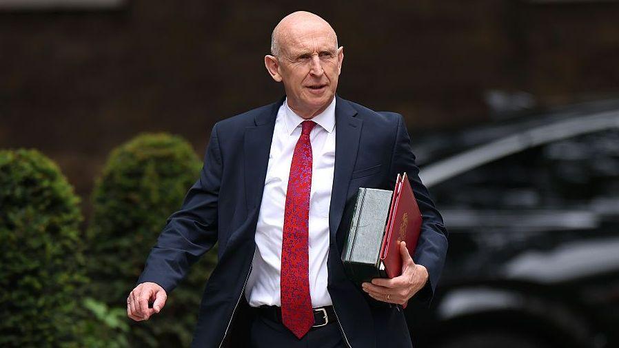 John Healey walks along Downing Street with files under his arm wearing a dark suit and a long red tie.