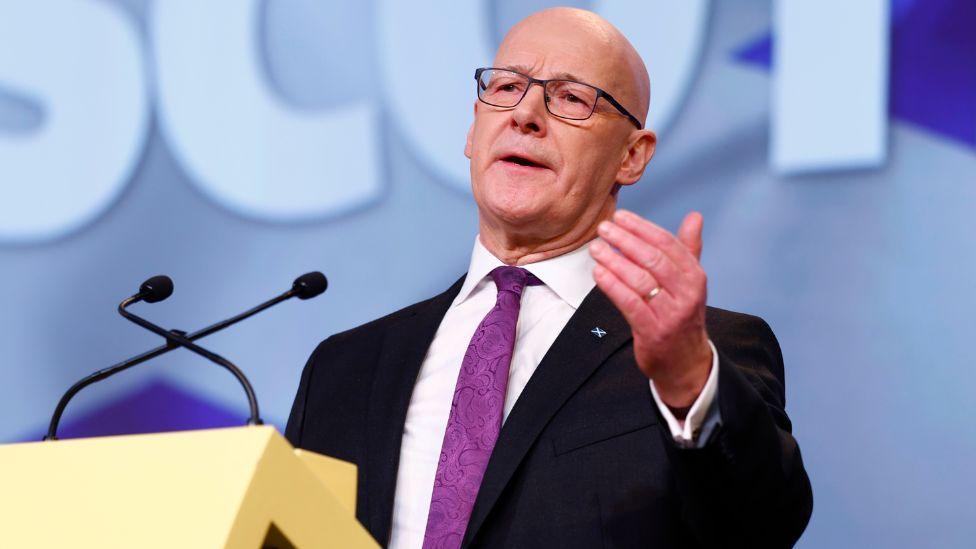 John Swinney, who is bald man with glasses, wearing a black suit and purple tie with a saltire pin on it. He has his left hand held in front of him