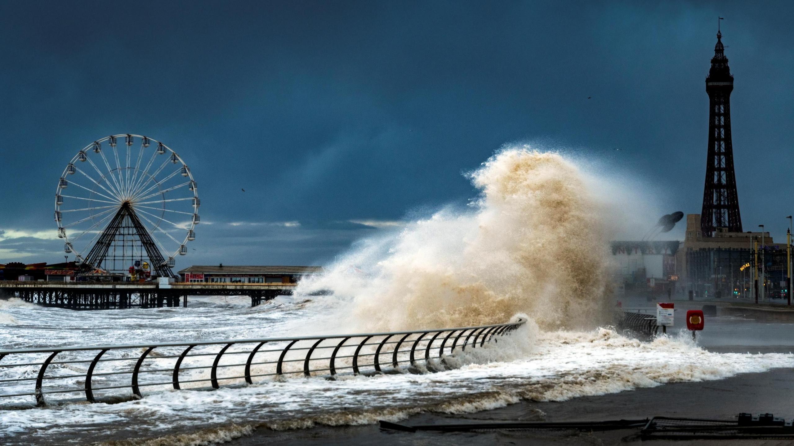 Large waves crash onto the promenade at Blackpool. Blackpool Tower and a ferris wheel are in the background.