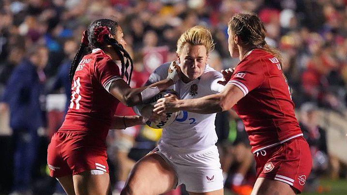 An England player, wearing white and carrying the ball in her right arm, is tackled by two Canadians, in red