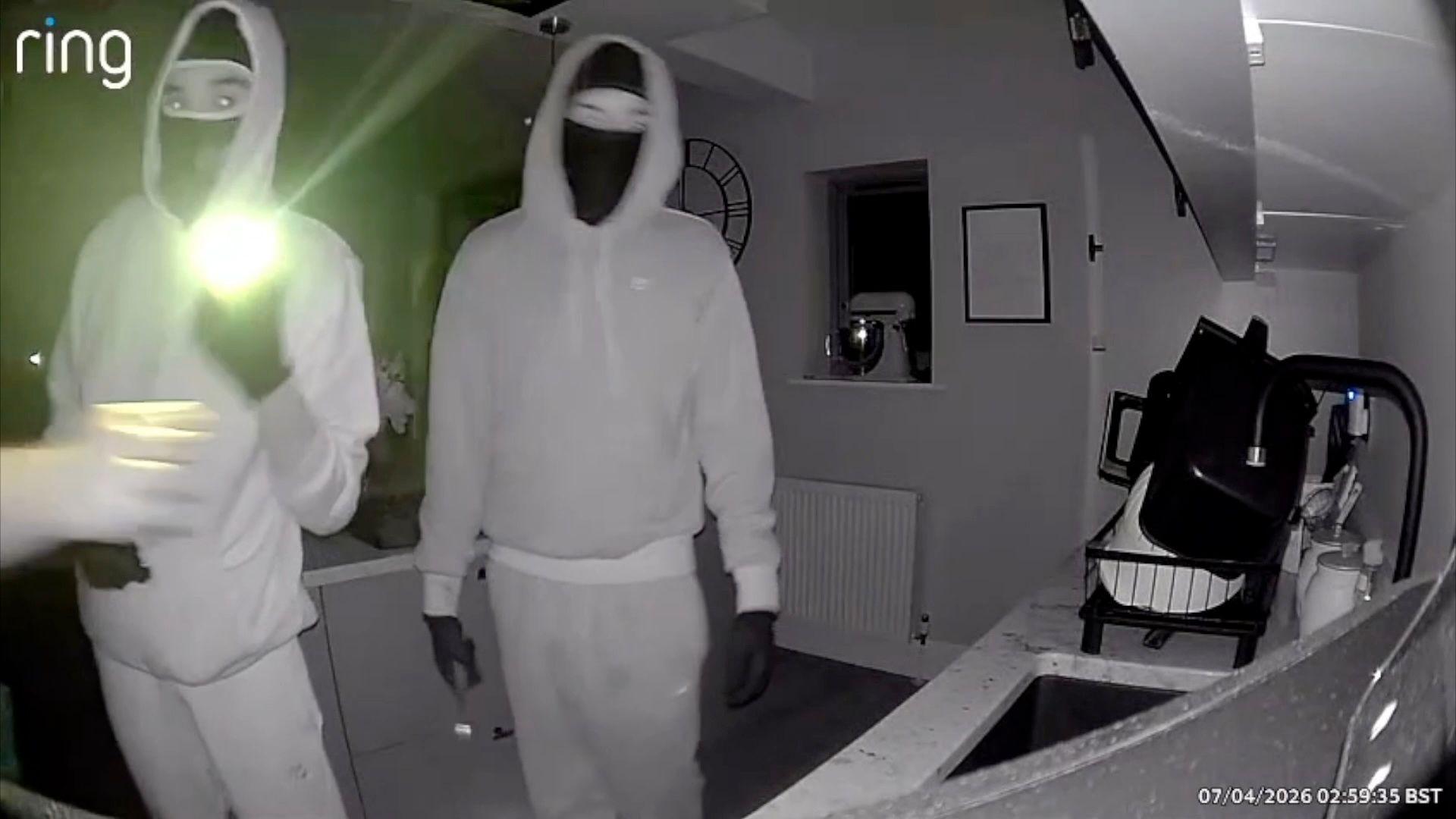 A still from black-and-white Ring camera footage of two burglars wearing light-coloured hoodies and dark balaclavas, one of them holding a torch, as they stand inside the kitchen of a home.
