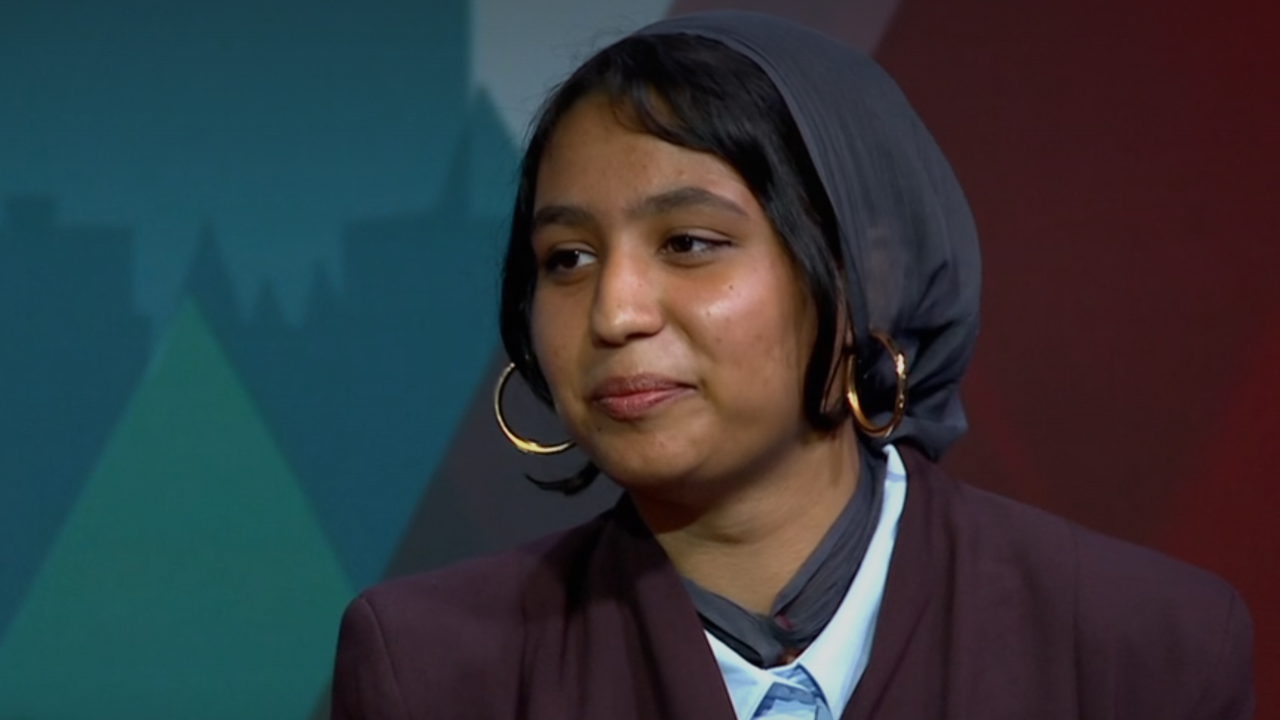 A head and shoulders image of a young woman wearing a purple blazer over a light blue shirt, gold hoop earrings and a headscarf.