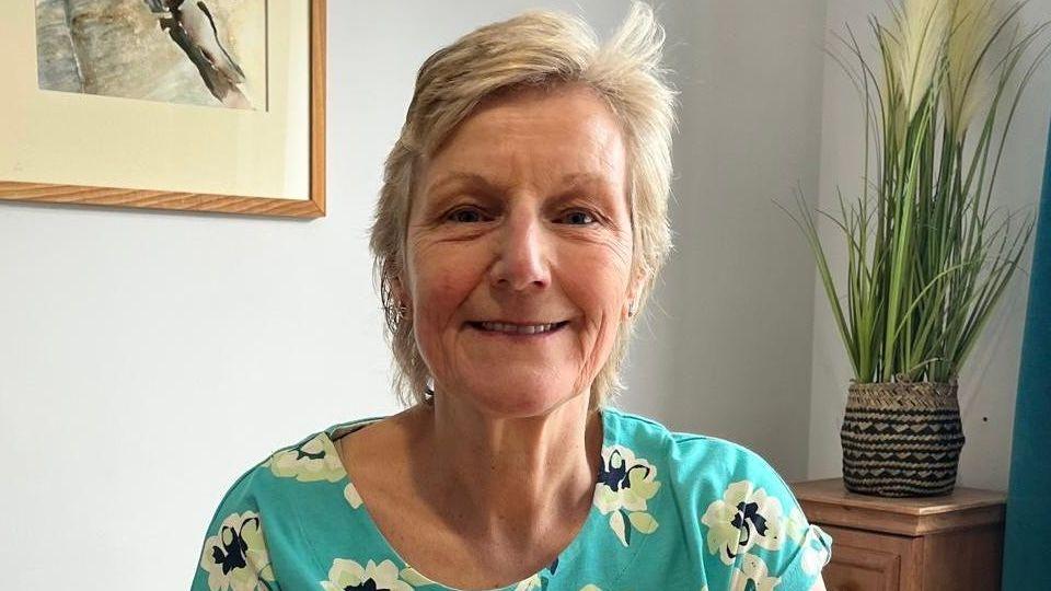 Head and shoulders shot of Sue Lawes, a middle aged woman in a blue flower-patterned top, in a living room