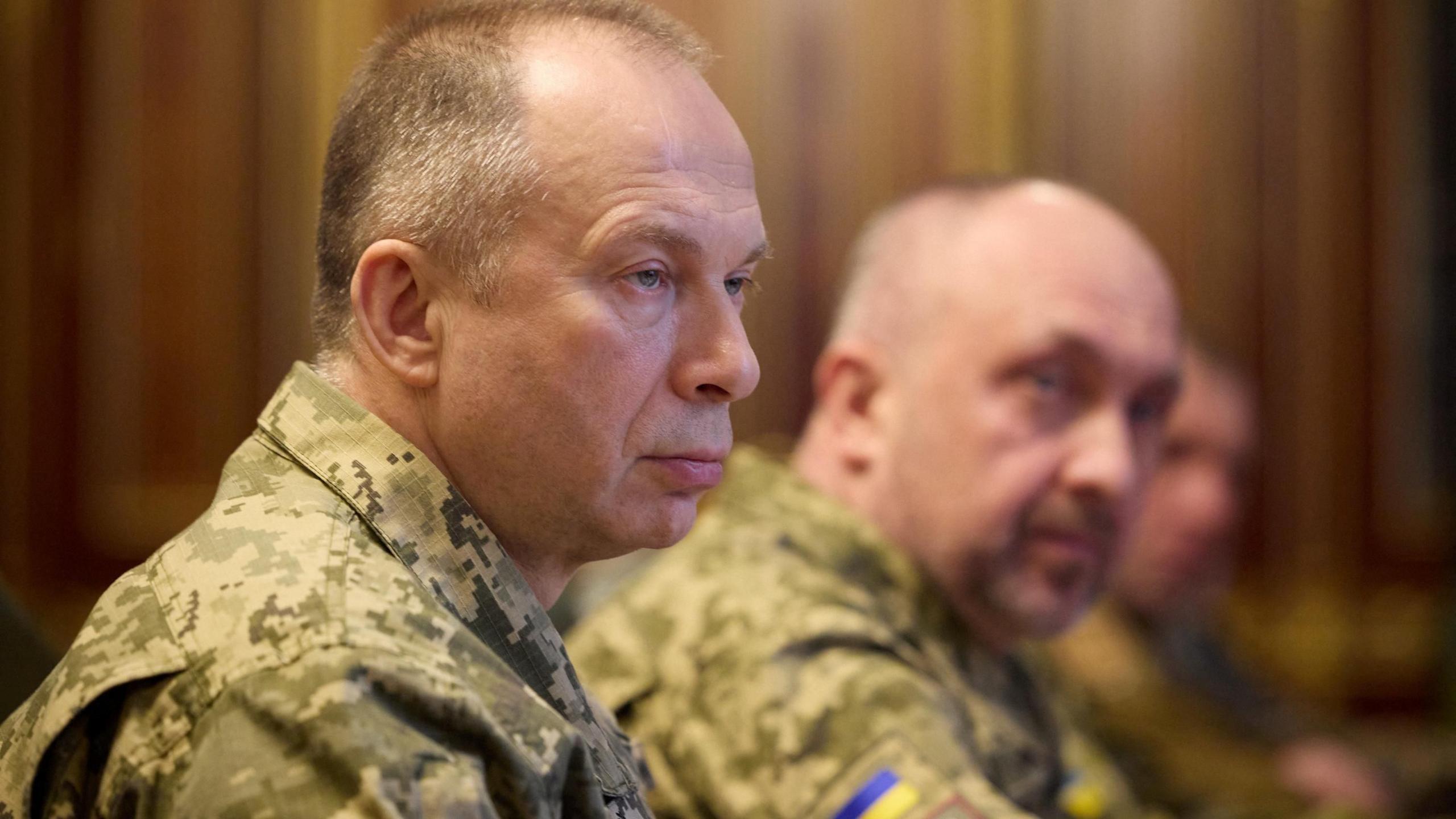Gen Syrskyi pictured from the side. He is wearing green military fatigues and has short grey hair. He is sat beside two other men in the same uniforms, with the blue-and-yellow Ukraine flag on their shoulders. They are sat in front of a wall of wooden panels.