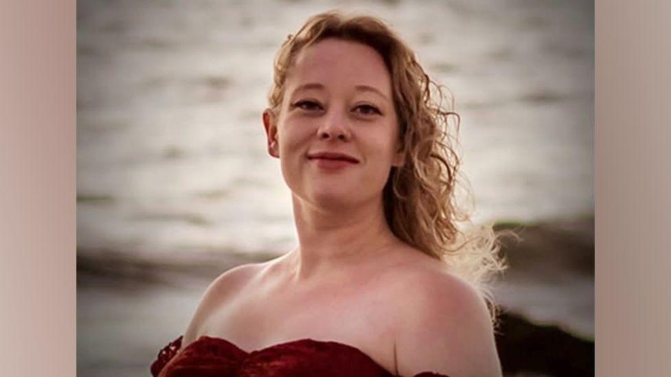 Renee Good has long, blonde, curly hair and is wearing a red off-the-shoulder top. The ocean is in the background.