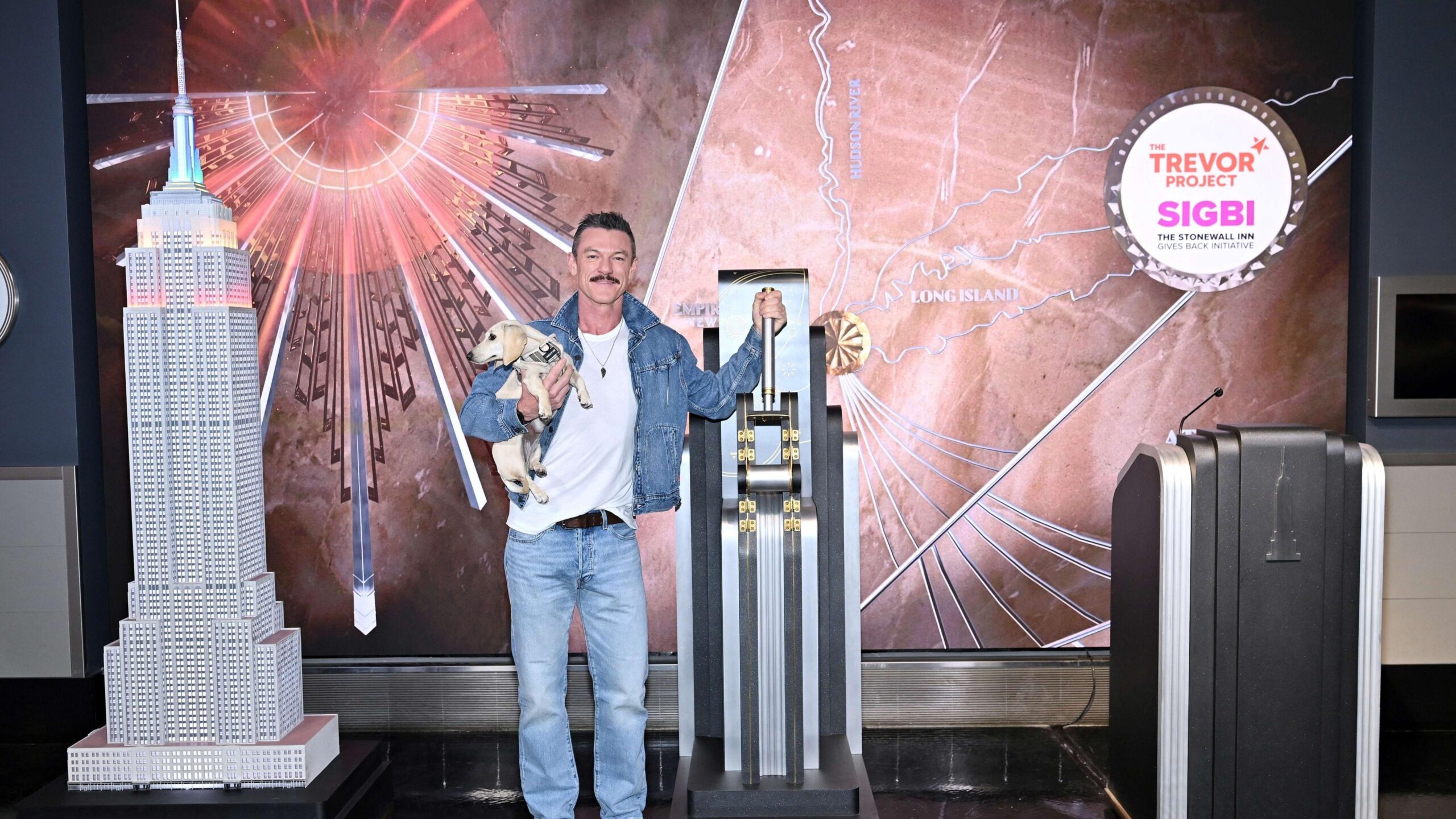 Luke Evans wearing double denim and a white top with his hand on a leaver. He's also holding on to his cream miniature dachshund.