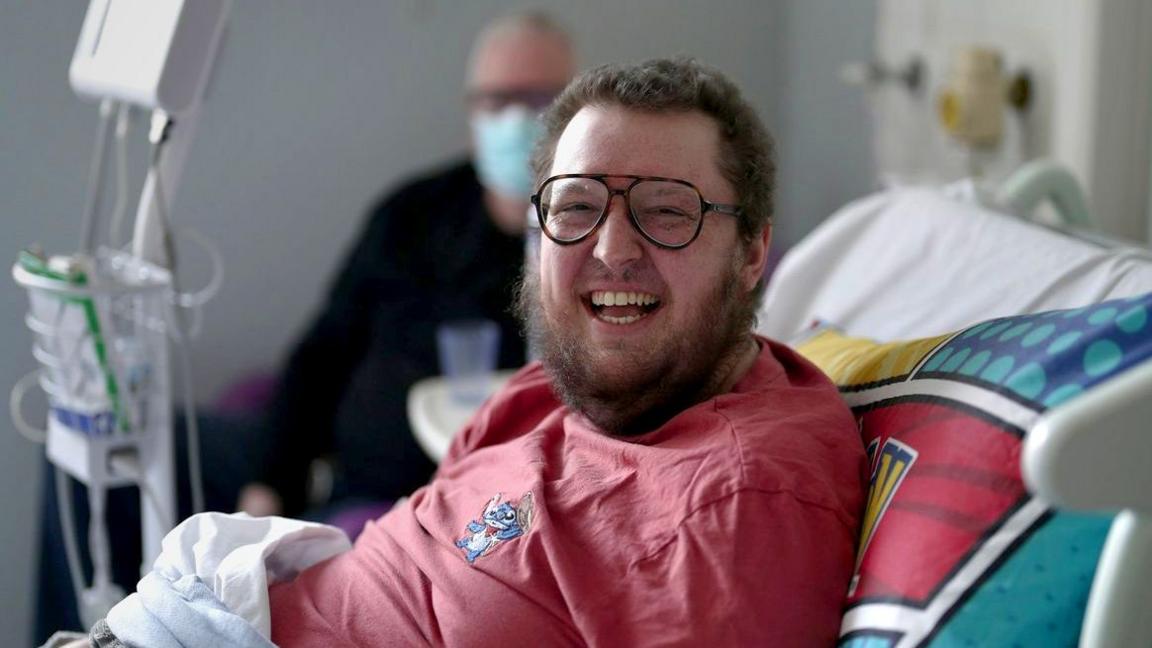 A man with brown hair and a beard smiling wearing aviator-style glasses sits in a hospital bed. He is wearing a red Lilo and Stitch top