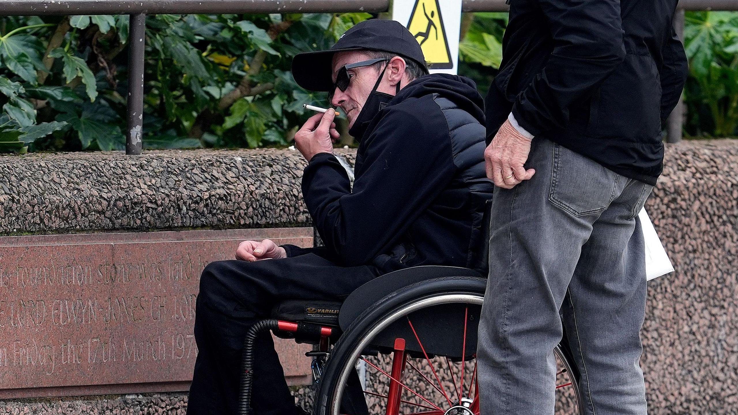 A man in a wheelchair wearing a dark navy hooded jacket with a quilted front is photographed holding a ciggarette towards his mouth. He is wearing dark sunglasses.