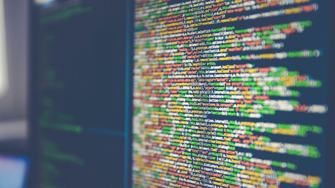 A close-up photograph shows a computer monitor filled with colourful programming code. Lines of green, yellow, orange and white text stand out against a dark blue background. The screen is photographed at an angle, with the centre sharper and the edges blurred. Another dark monitor sits to the far left.