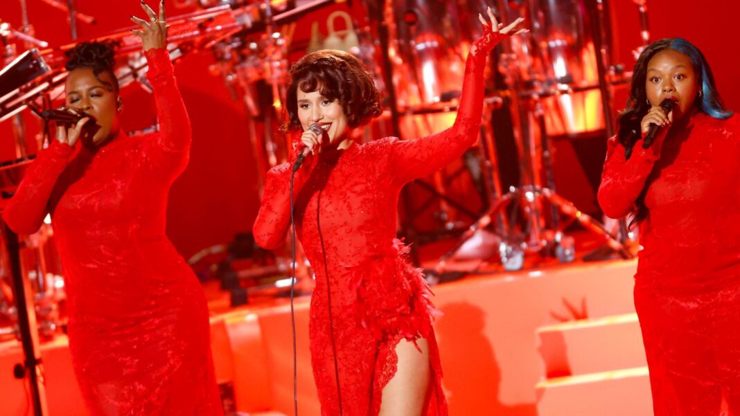 Raye sings into a microphone while performing on stage. She wears a red lace dress with long-sleeves and a high leg split. There are back-up singers performing beside her and a band in the background.
