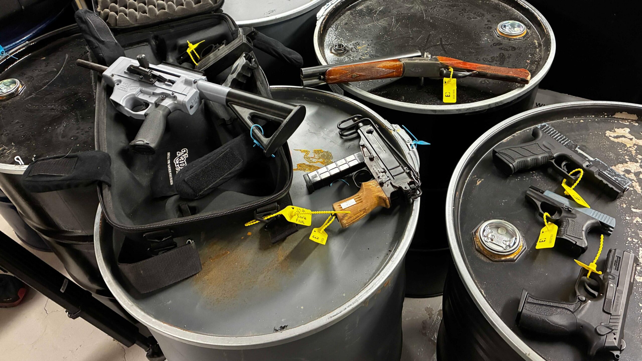 A selection of guns with yellow tags are displayed on top of some silver cylinders. There is a shotgun with brown handles, a grey 3D printed semi automatic gun, a silver firearm and three black firearms.