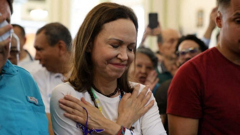 A woman in white crosses her arms on her chest with her hands pointed towards her shoulders and her eyes closed