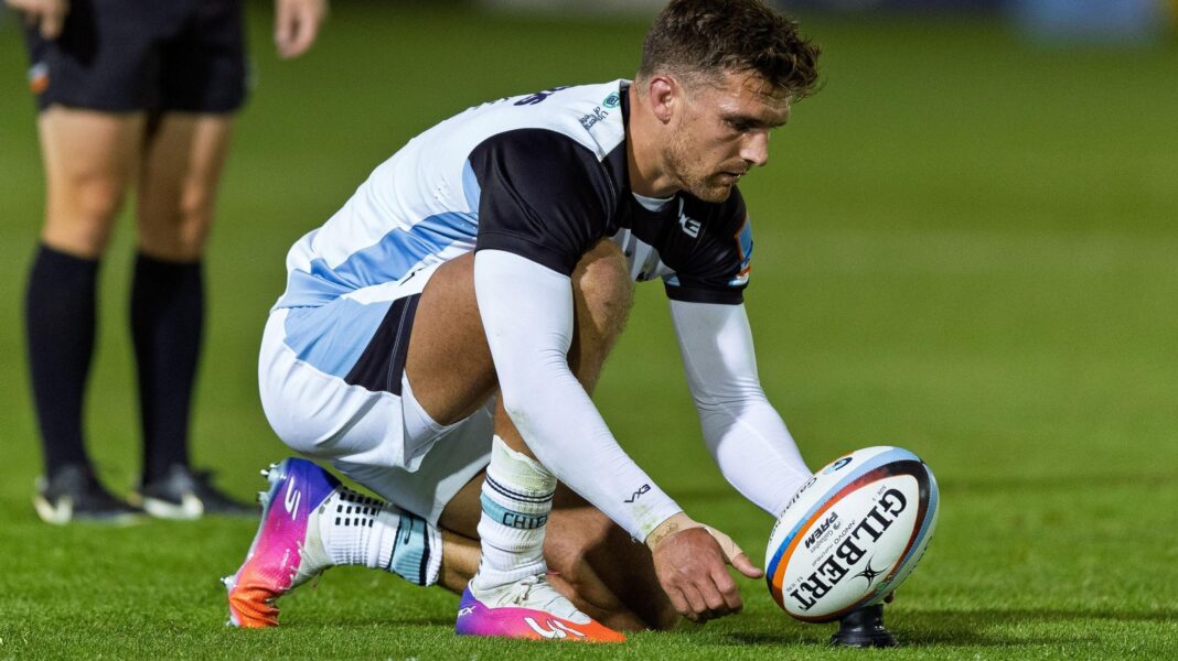 Henry Slade kneels on the grass on the pitch and holds his hand towards a rugby ball that is resting on a tee, as he prepares to take a kick at the posts