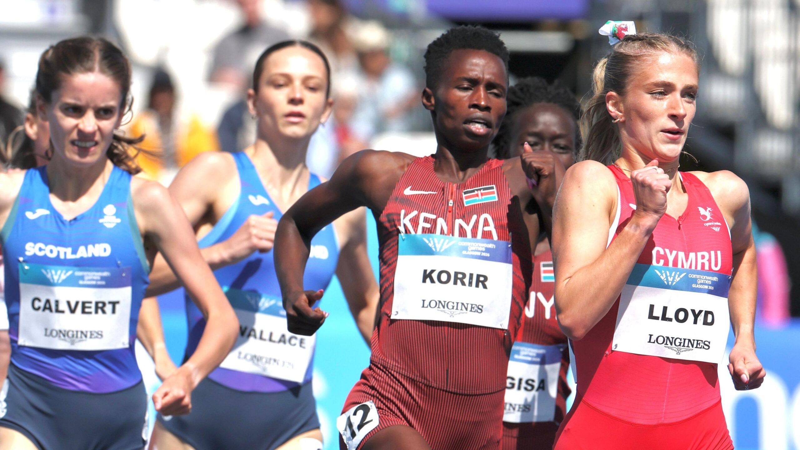 Naomi Korir visible from thigh-level runs in the centre of a pack, moving left to right, during a mile race at the 2026 Commonwealth Games in Glasgow. Korir, who is wearing a maroon Kenyan zip-up running top and shorts with a white name bib on her upper stomach, has a look of deep concentration on her face and is pumping her arms hard. A Welsh athlete in red running kit leads Korir, while two Scottish runners in blue kit trail behind. Blurred in the background is a crowd sitting in the sun