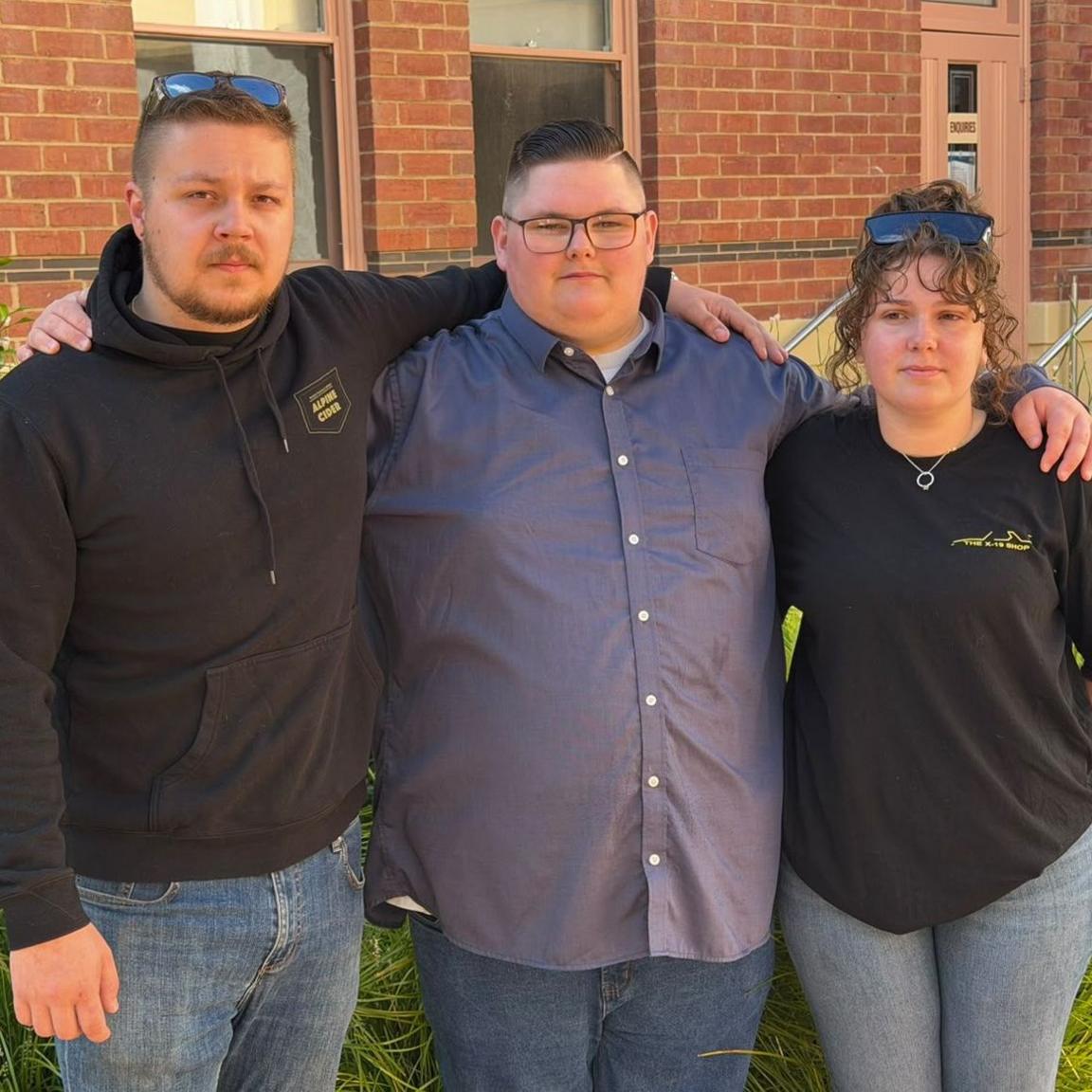 Three people wearing dark clothing and their arms around each other pose for photo