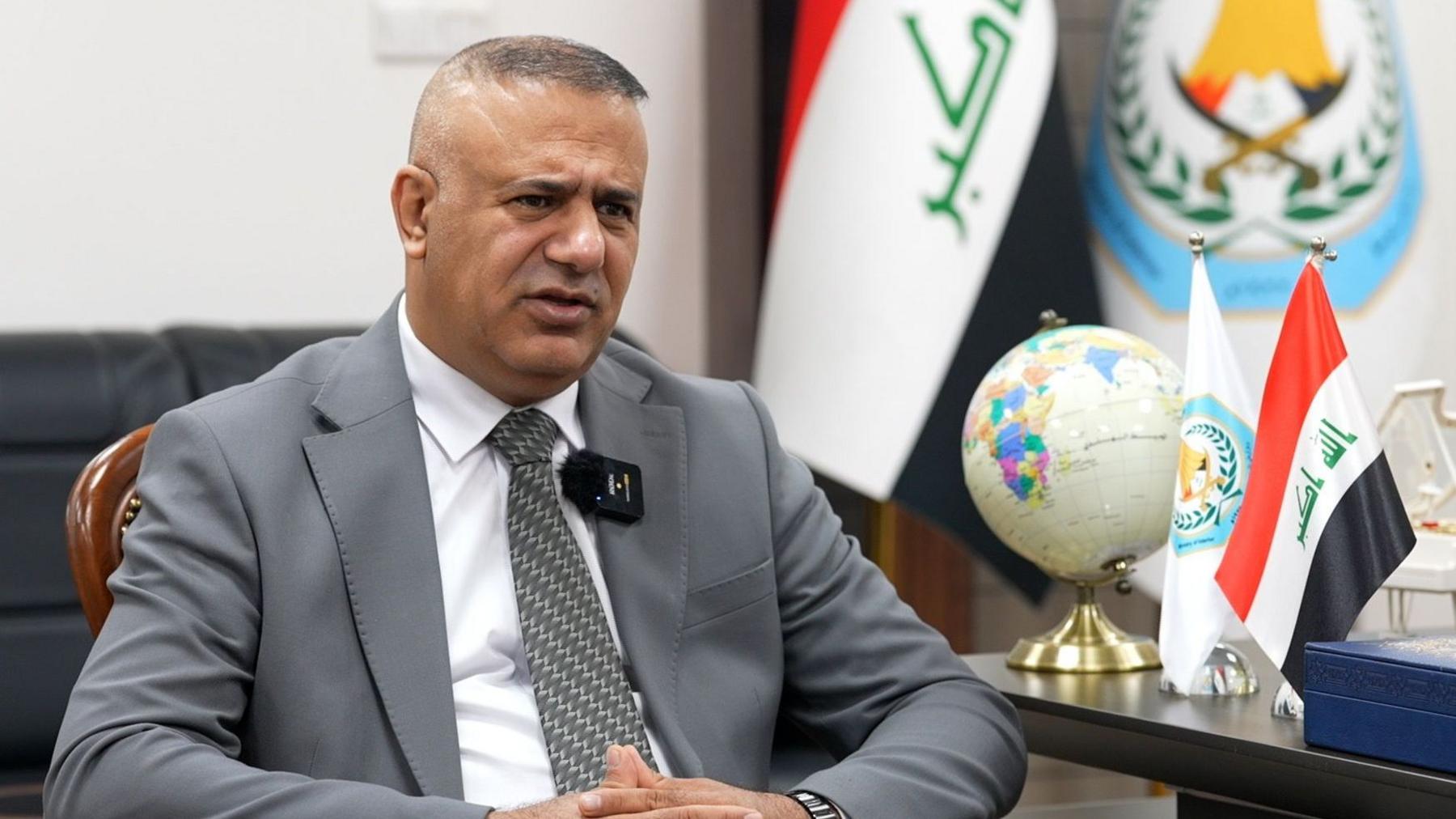 Muqdad Miri in a grey jacket, white shirt and grey patterned tie, in an office next to Iraqi flags and a globe on table