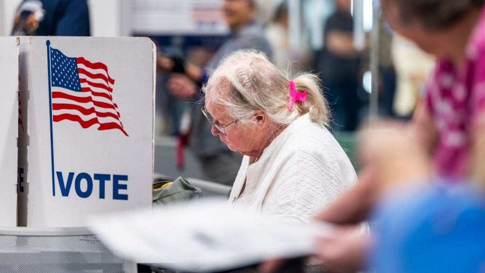 An older woman, wearing metal rimmed glasses, a white sweater, and with her white hair pulled up in a pink claw clip, sits in front of a voting booth. On it is a US flag and the word 'VOTE' in blue.