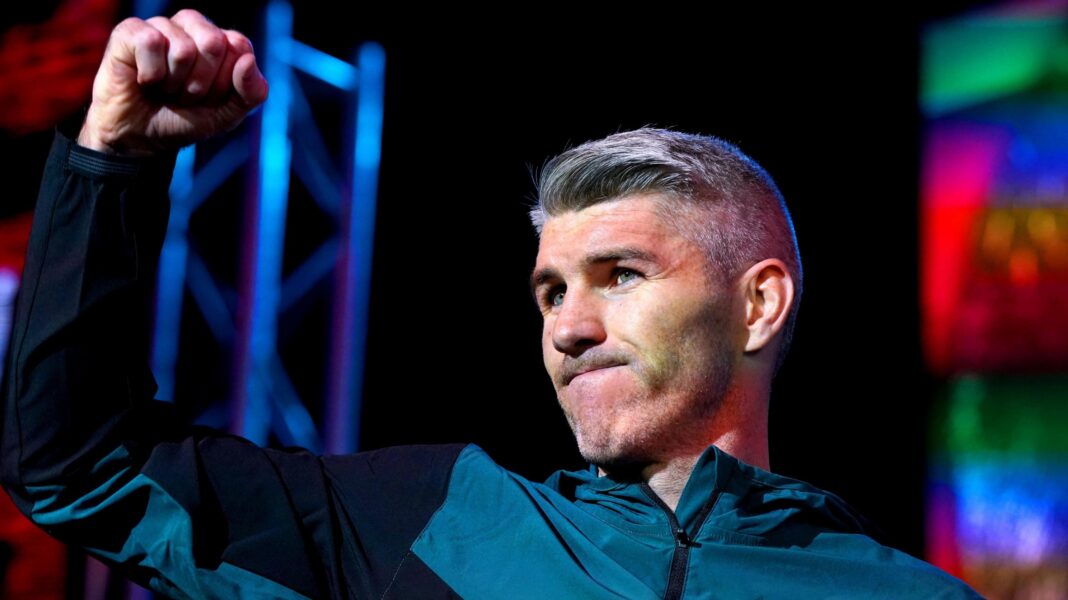 A man in his 30s, with grey hair and wearing a turquoise and blue tracksuit, stands with his right arm raised in the air and his hand clenched into a fist.