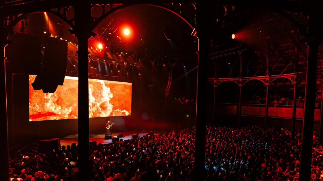 Anuv performing on stage with a guitar. The crowds are lit by the red lighting coming off the stage.