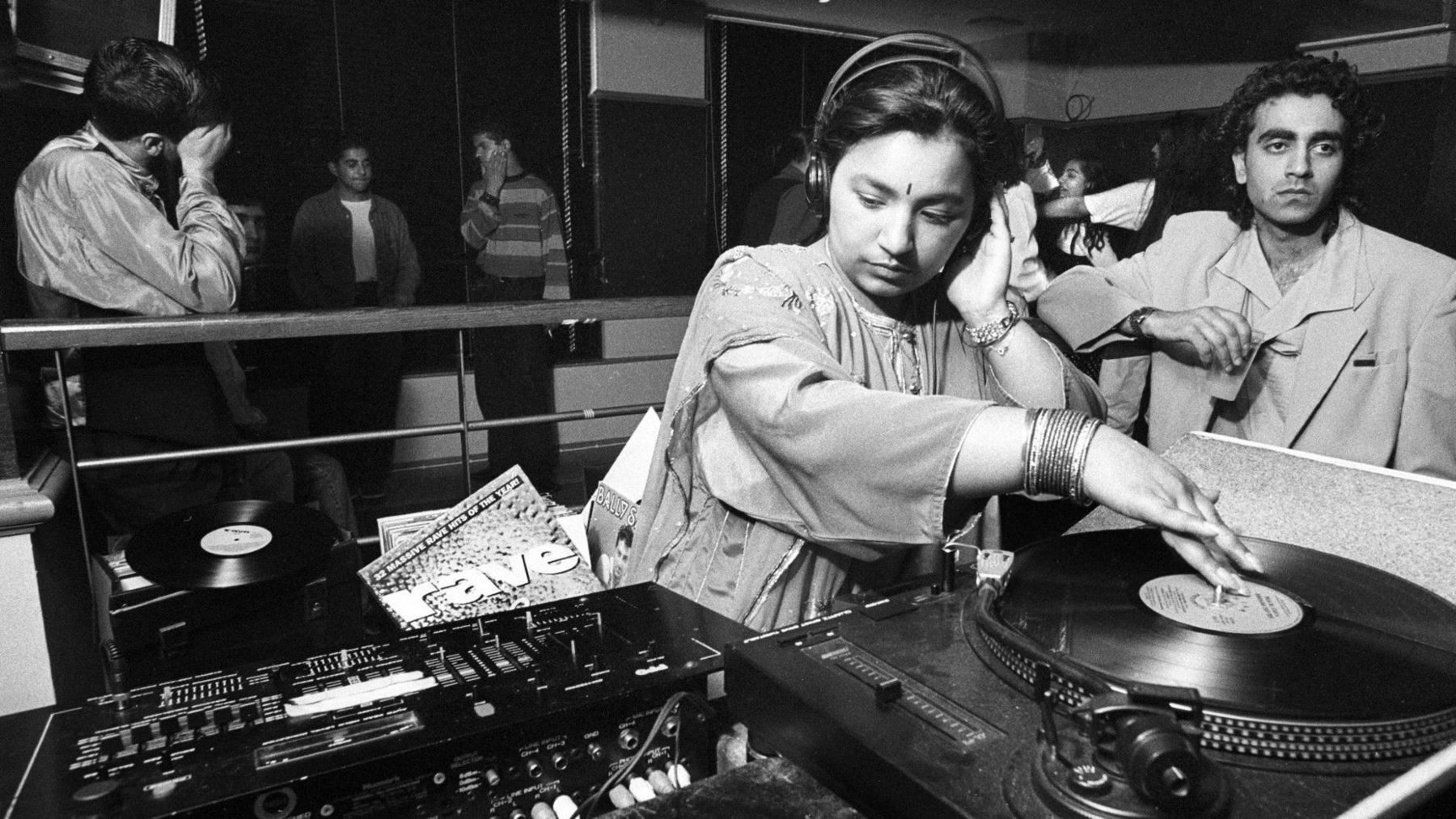 A black and white photo of a young woman spinning a record on DJ decks. She is wearing traditional South Asian clothing as well as a bindi and bangles. Groups of young people stand or dance in the background.
