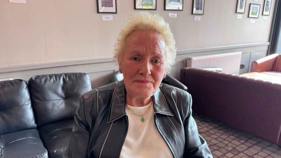 A woman with short blonde hair, wearing a black leather jacket and a white t-shirt