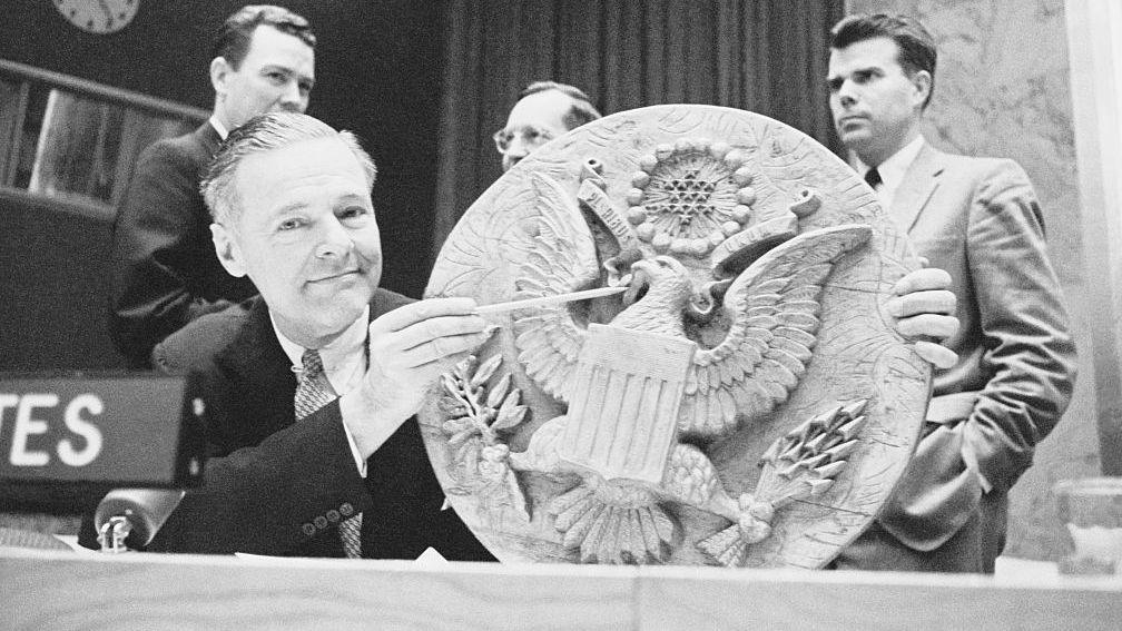 A black and white image of US Ambassador to the UN Henry Cabot displaying a wooden replica of the Great Seal of the United States, which Soviet boy scouts presented to US Ambassador Averell Harriman in 1945.