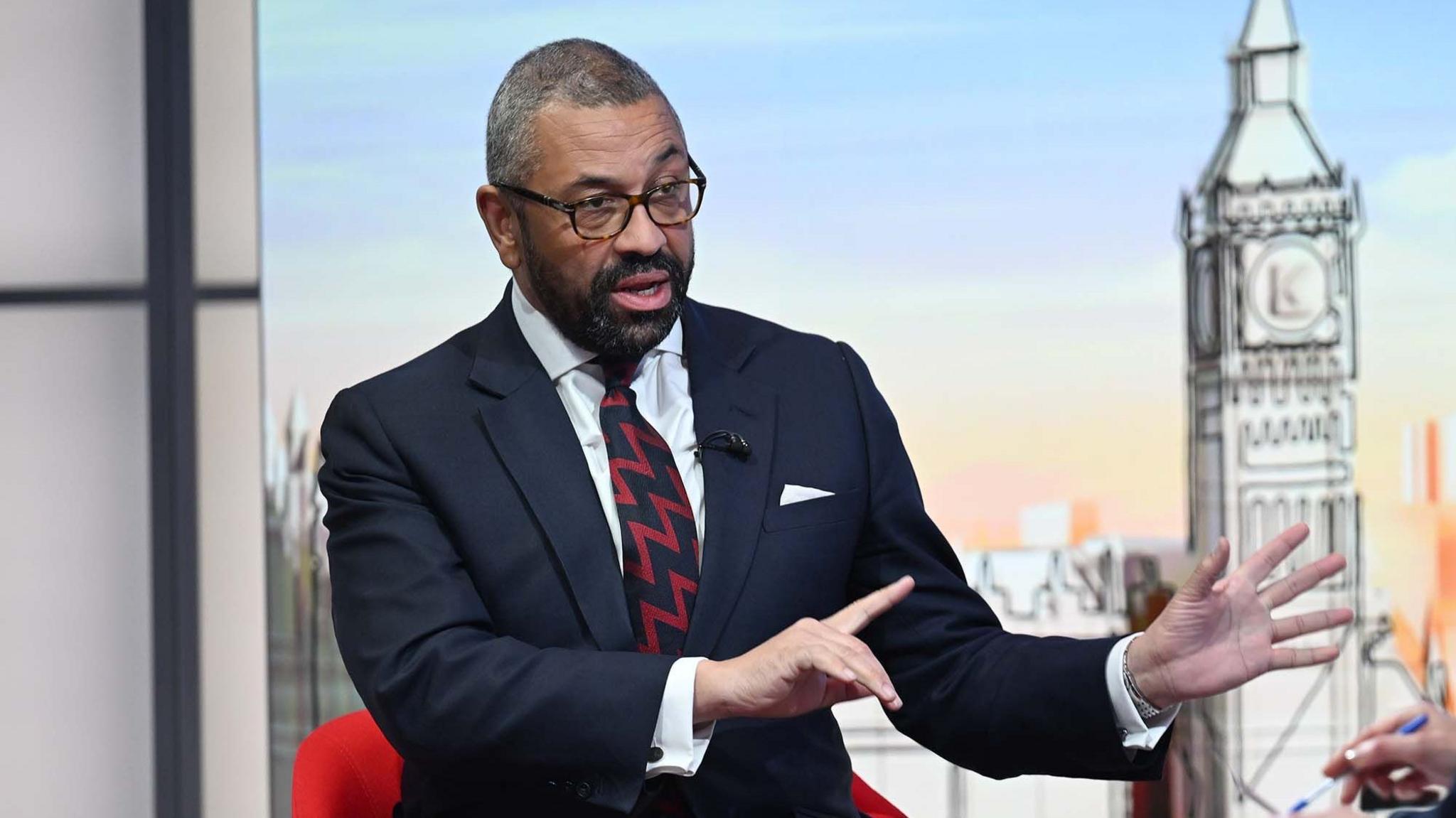 A middle aged man with dark hair and a short dark beard, being interviewed in a TV studio, he is wearing a dark blue suit white shirt and black and red patterned tie.