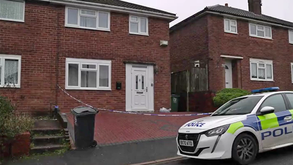 A police car is parked on the road outside a house, where police crime tape can be seen cordoning off the brick-paved driveway. The house has a white door and a white-framed front lower window.