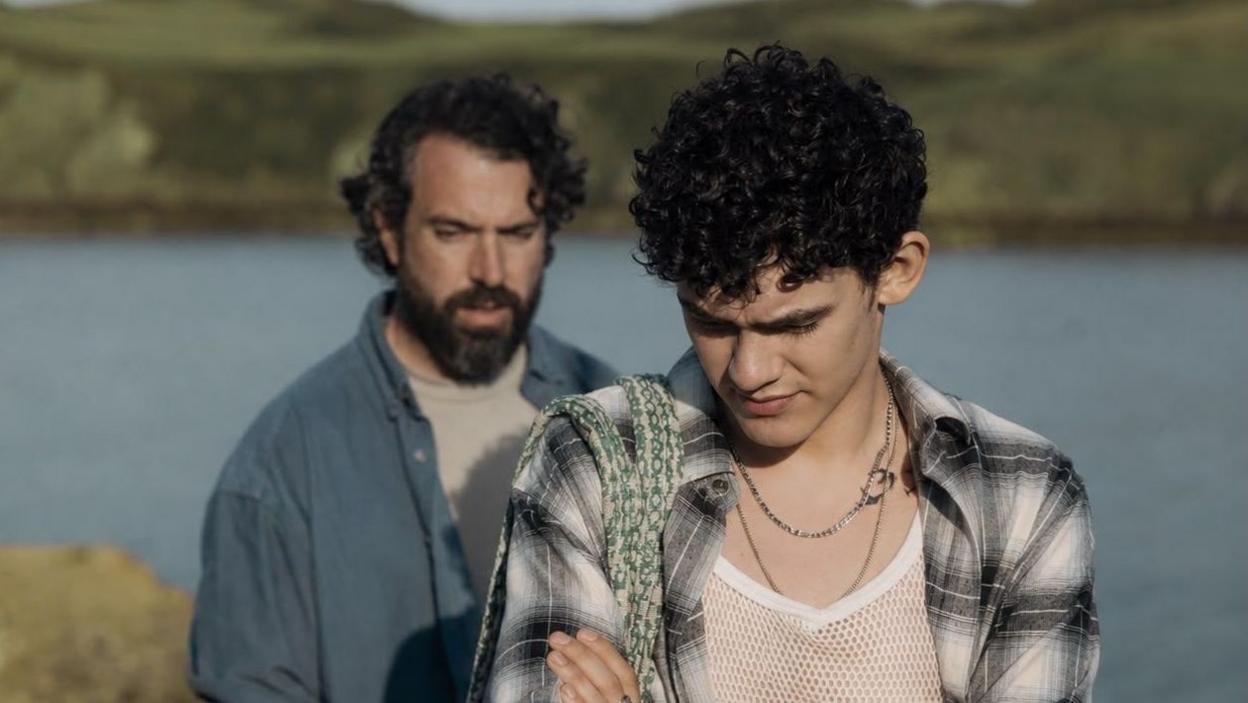 Tom Cullen and Joe Locke by the sea. Tom is behind Joe in a blue shirt and grey t-shirt, and has curly black hair. Joe is in front with curly black hair, a string vest and black and white checked top. Joe has his arms crossed and looks down.